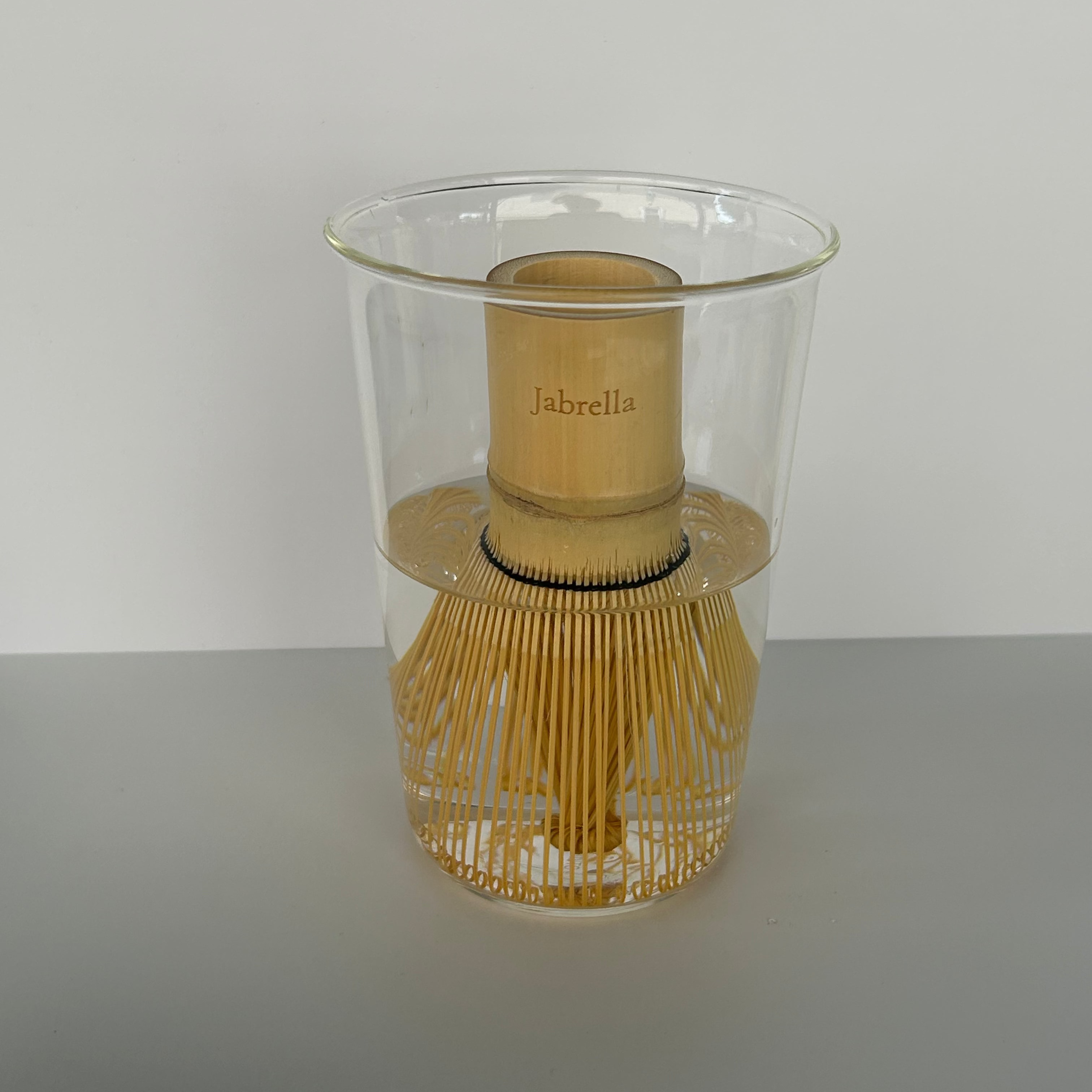 A clear glass cup with a bamboo whisk inside, branded 'Jabrella' on the bamboo collar, set against a plain light gray background.